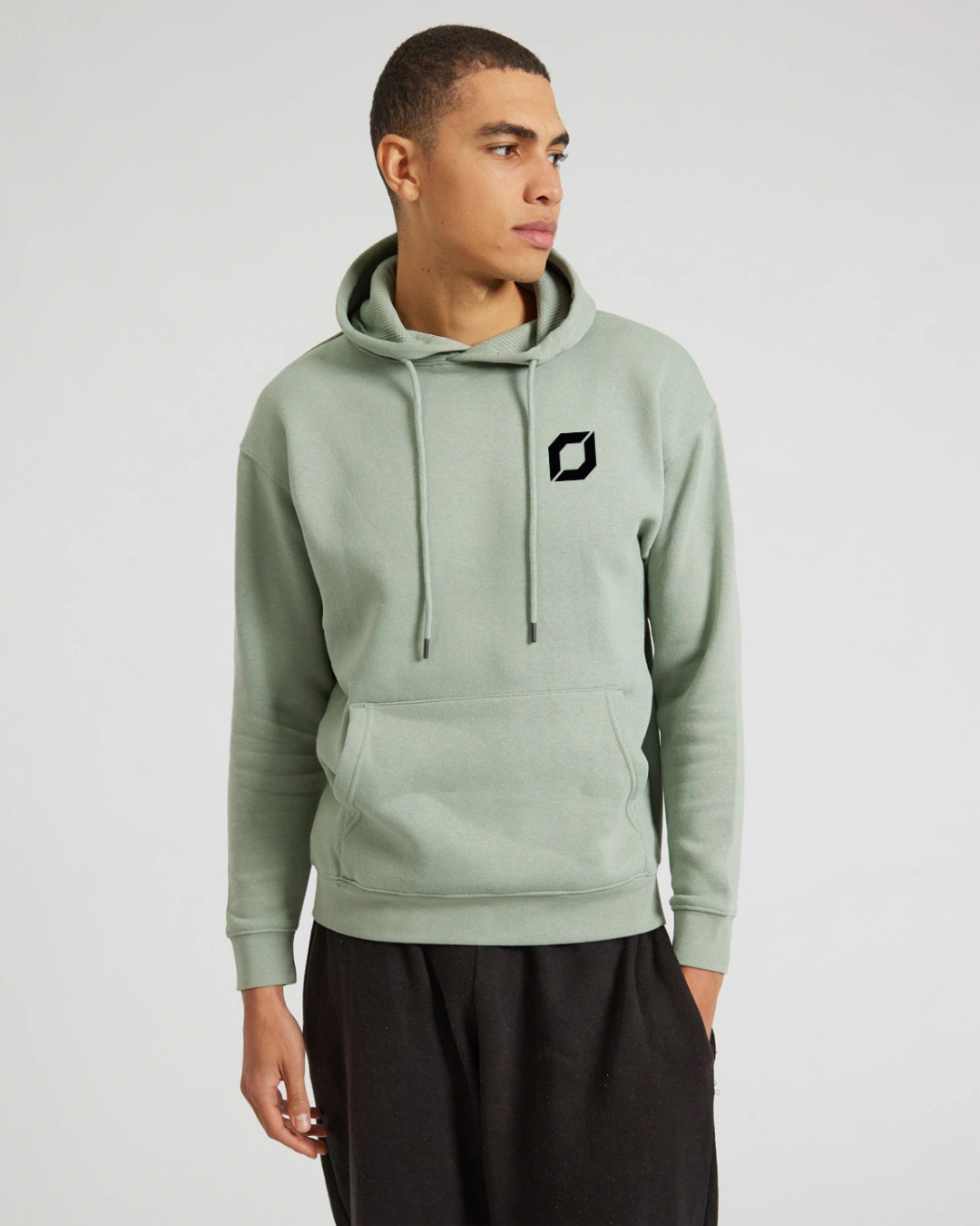 A man wearing a sage green pullover hoodie with a black obsidian logo on the left chest, paired with black sweatpants, photographed against a light gray studio background.