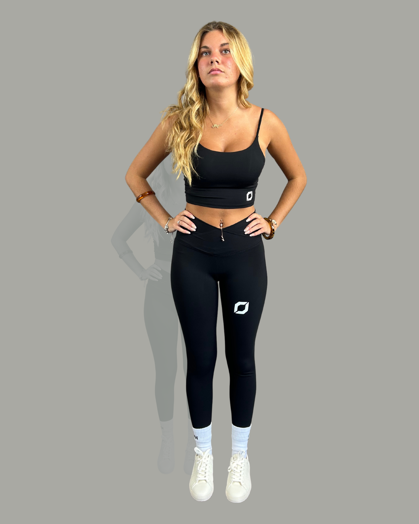A blonde woman modelling a matching black activewear set consisting of a crop top and high-waisted leggings, both featuring a white obsidian logo on the front and hip, paired with white socks and white sneakers, set against a solid grey background.